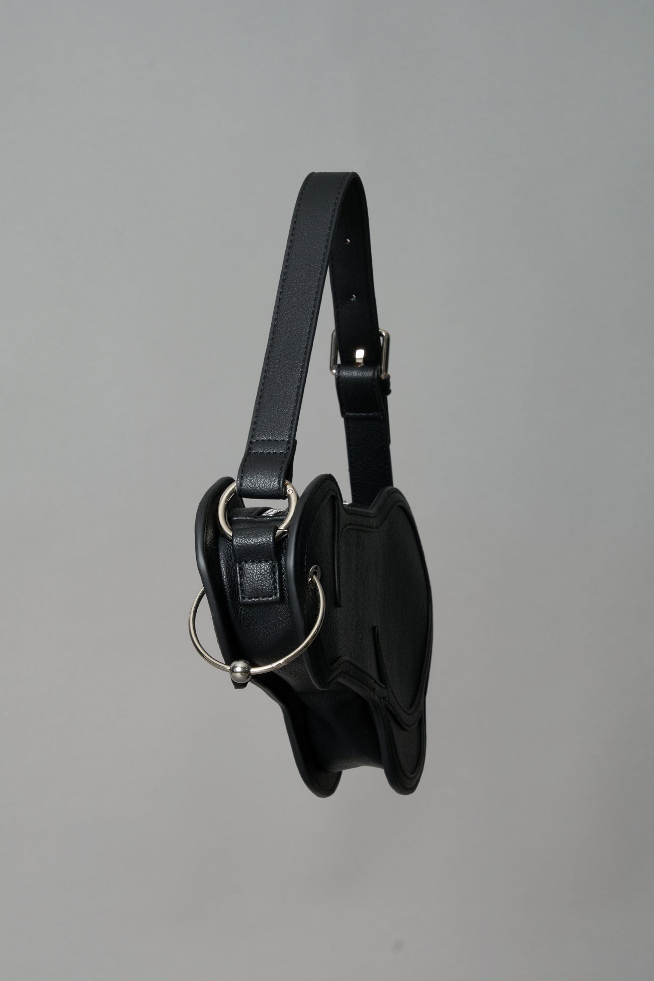 Black vegan leather shoulder bag, featuring a silver piercing detail and an adjustable strap, displayed against a neutral grey background."