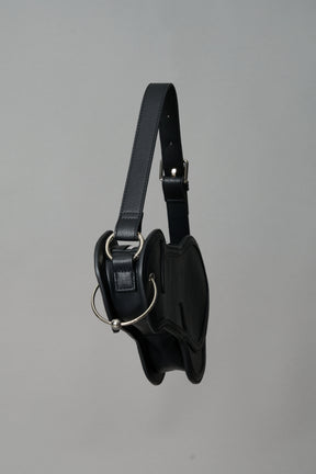 Black vegan leather shoulder bag, featuring a silver piercing detail and an adjustable strap, displayed against a neutral grey background."