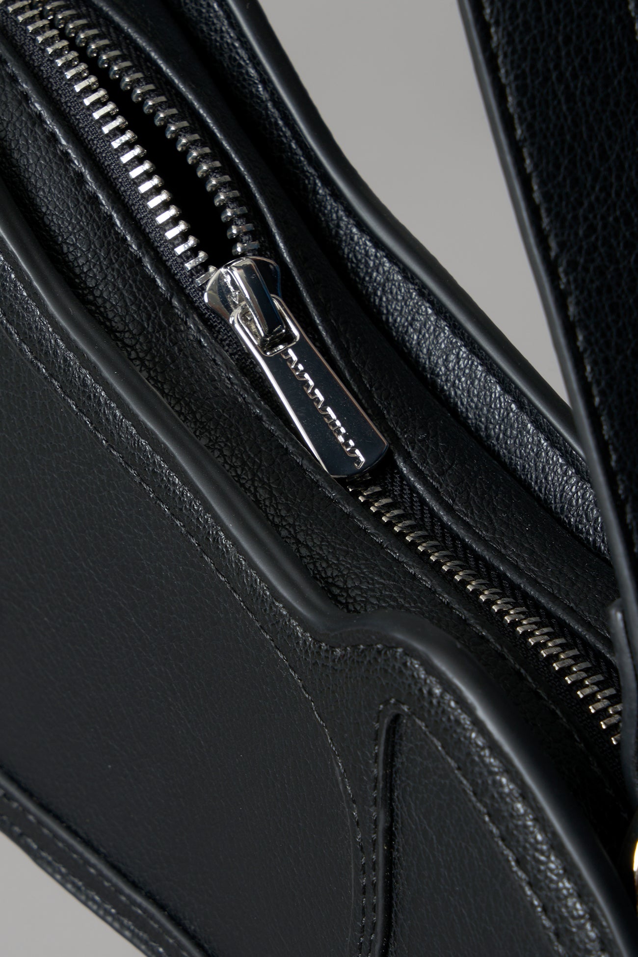 Black vegan leather shoulder bag, featuring a silver piercing detail and an adjustable strap, displayed against a neutral grey background."
