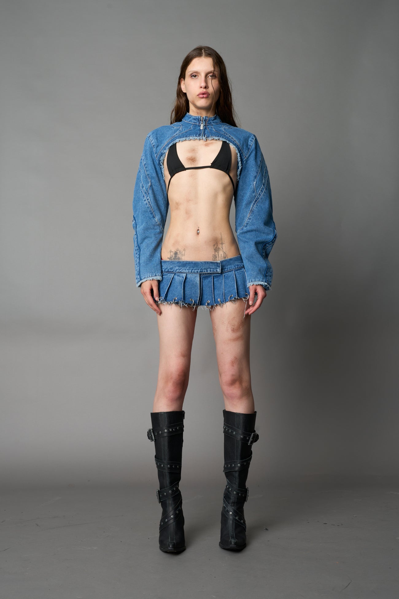 Model in ultra-cropped blue washed denim bolero jacket with front zipper, raw frayed edges, choker neckline, and sculpted biker sleeves