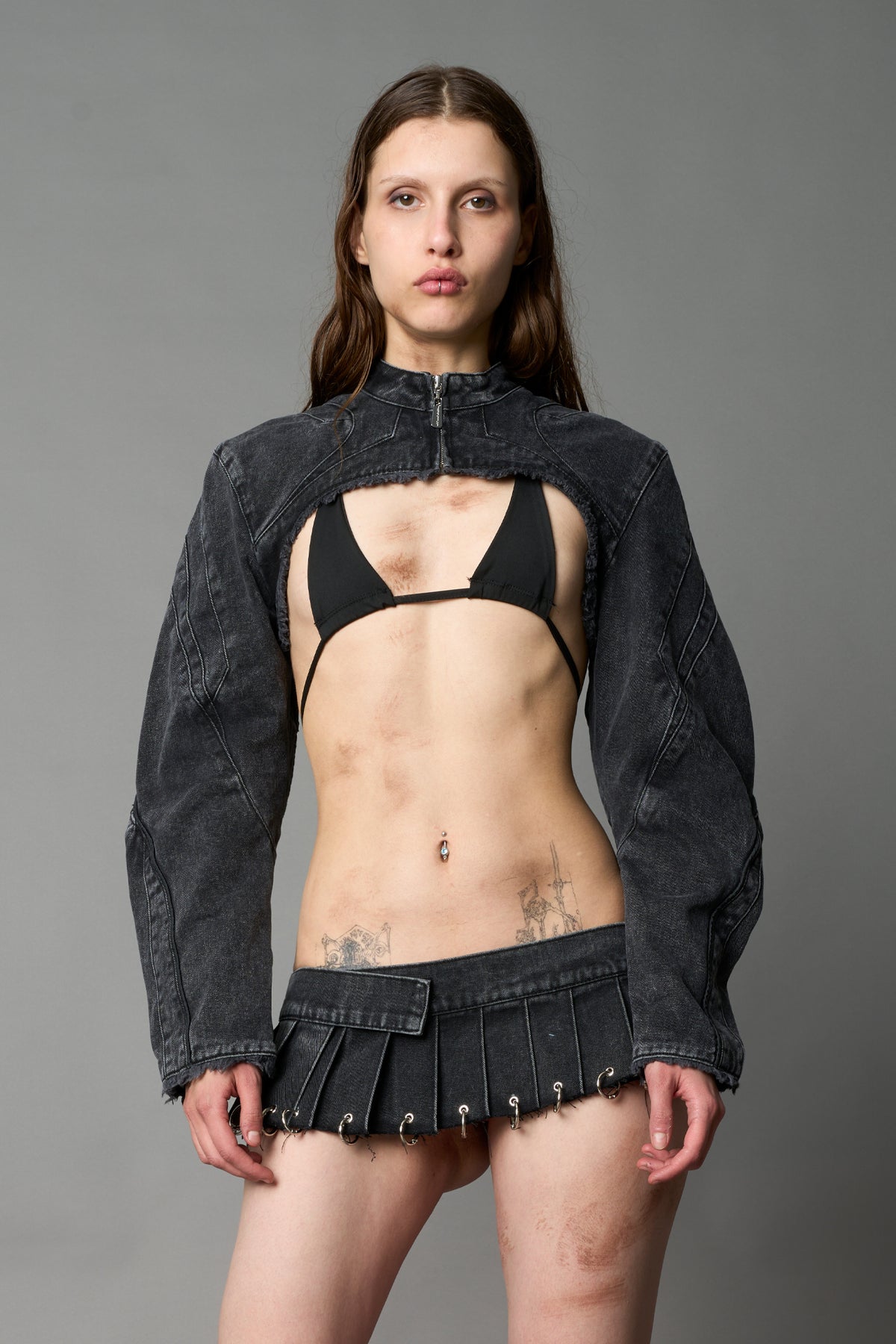 Model in ultra-cropped washed black denim bolero jacket with front zipper, raw frayed edges, choker neckline, and sculpted biker sleeves