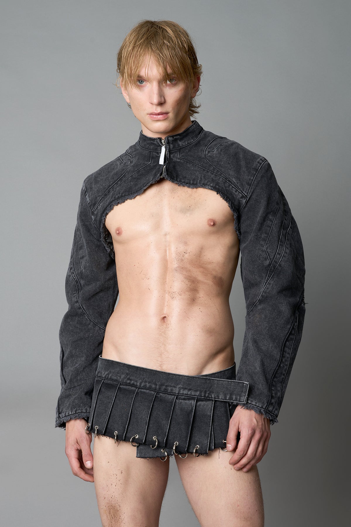 Model in ultra-cropped washed black denim bolero jacket with front zipper, raw frayed edges, choker neckline, and sculpted biker sleeves