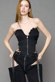 Black dark washed denim cone corset by NAMILIA with spiral-stitched cone cups, strapless fitted silhouette, front zip closure, lace-up sides, and raw frayed edges, worn by model on neutral background.
