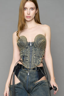 blue mud washed denim cone corset by NAMILIA with spiral-stitched cone cups, strapless fitted silhouette, front zip closure, lace-up sides, and raw frayed edges, worn by model on neutral background.