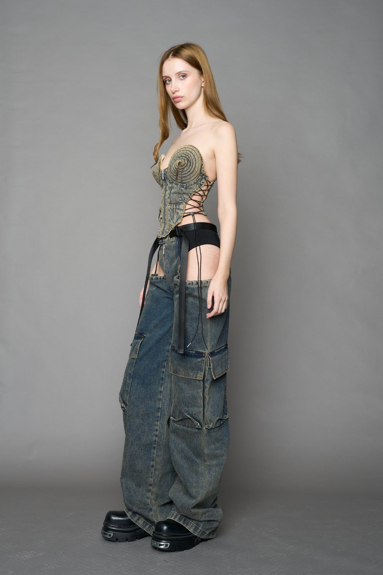 blue mud washed denim cone corset by NAMILIA with spiral-stitched cone cups, strapless fitted silhouette, front zip closure, lace-up sides, and raw frayed edges, worn by model on neutral background.