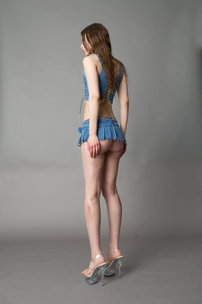 model in ultra-short blue washed denim pleated mini skirt featuring frayed hem, silver grommet rings, and wide belt-style front closure