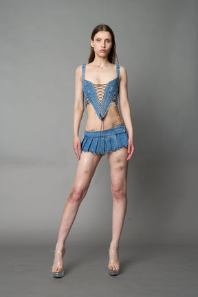 model in ultra-short blue washed denim pleated mini skirt featuring frayed hem, silver grommet rings, and wide belt-style front closure