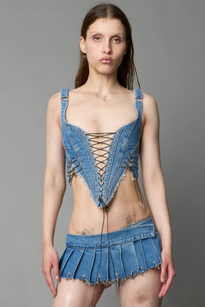 Model wearing blue washed denim corset top with plunging neckline, lace-up front and sides, raw frayed hem, and structured fit