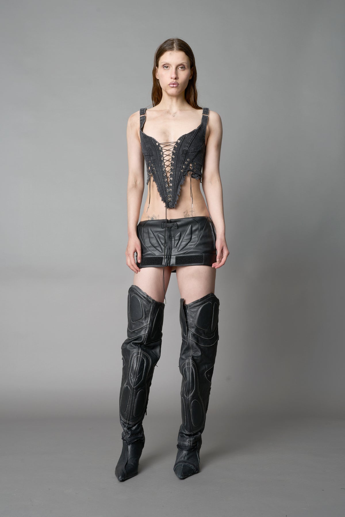 Model wearing washed black denim corset top with plunging neckline, lace-up front and sides, raw frayed hem, and structured fit