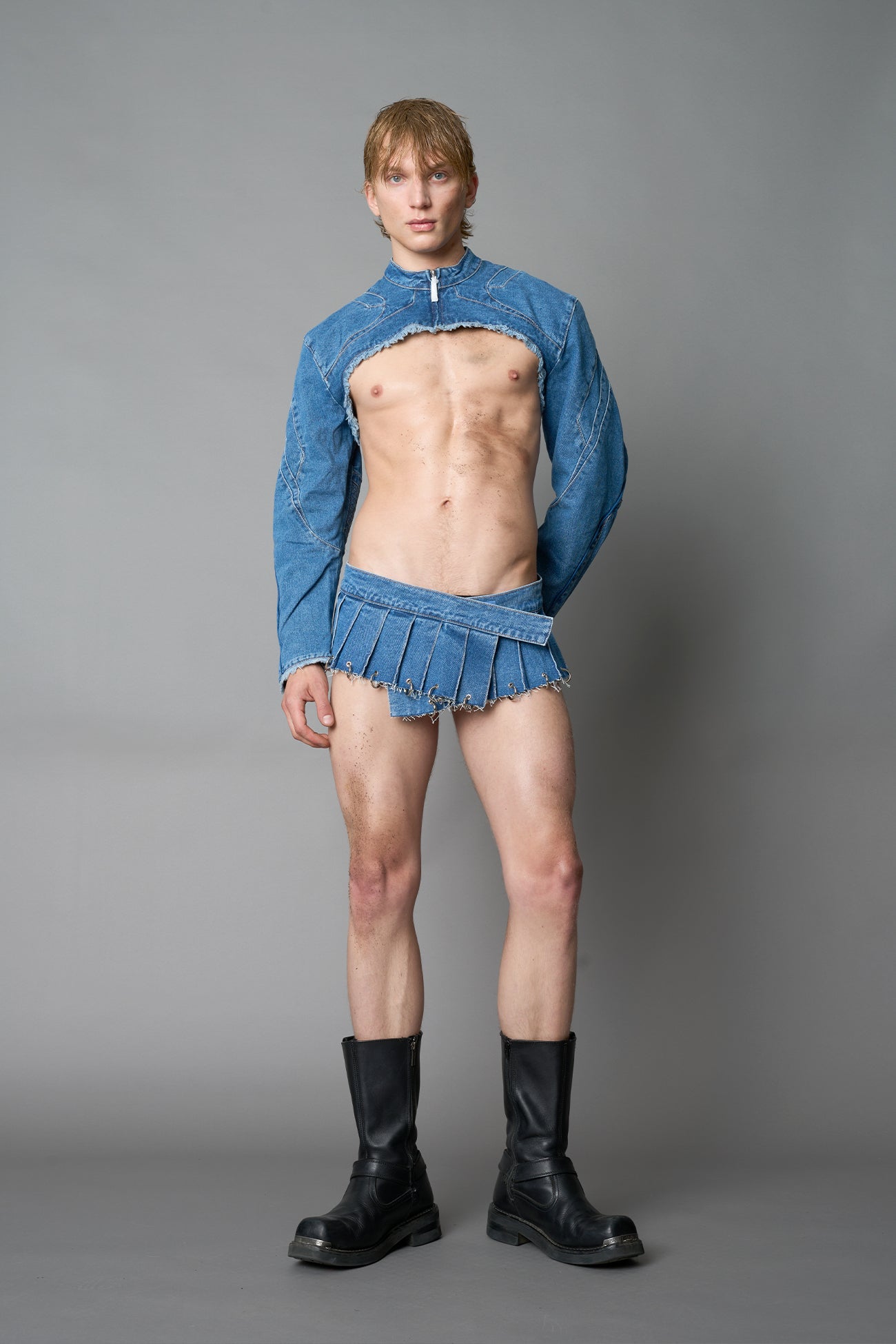 Model in ultra-cropped blue washed denim bolero jacket with front zipper, raw frayed edges, choker neckline, and sculpted biker sleeves