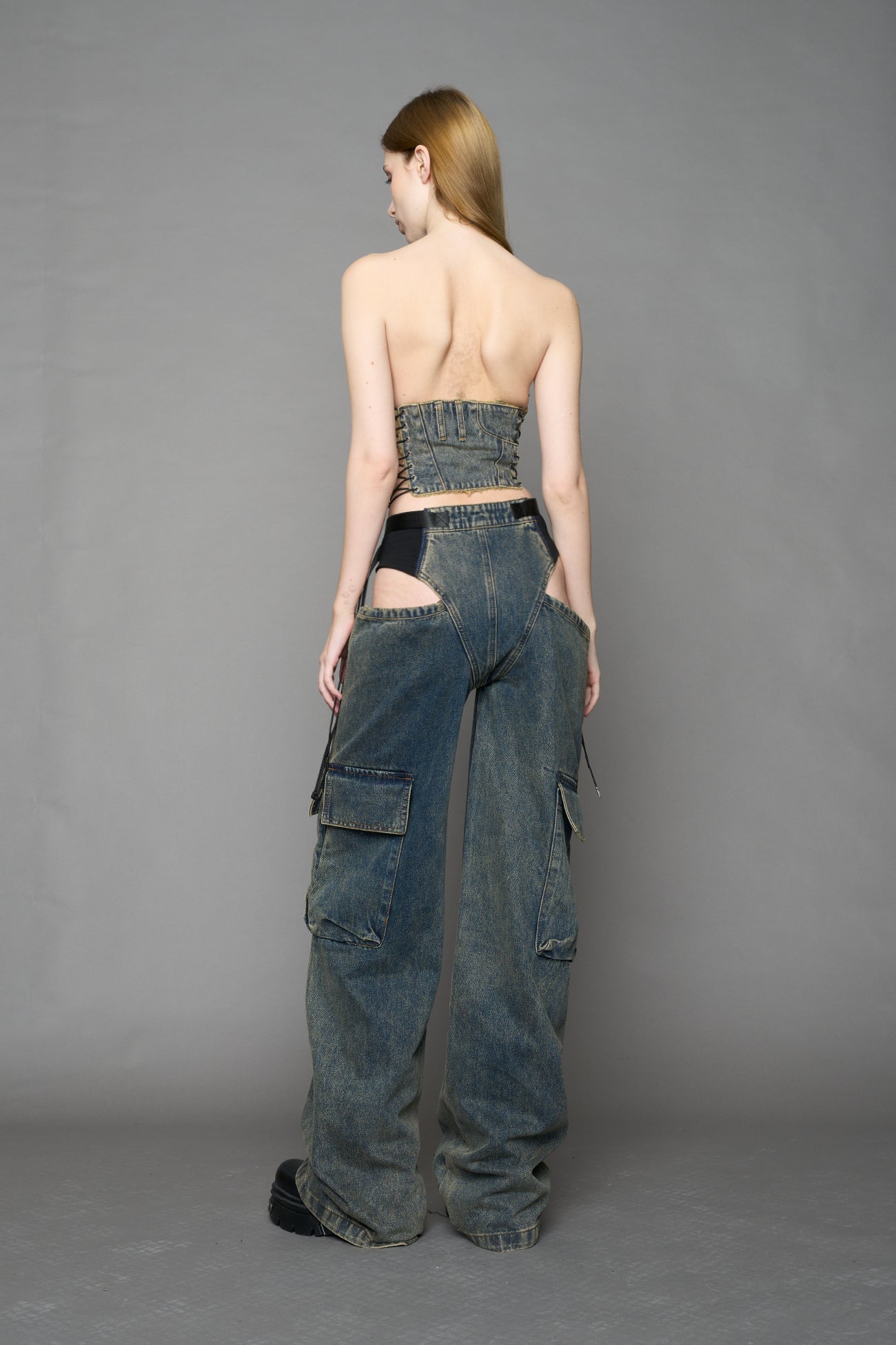 blue mud washed denim cone corset by NAMILIA with spiral-stitched cone cups, strapless fitted silhouette, front zip closure, lace-up sides, and raw frayed edges, worn by model on neutral background.