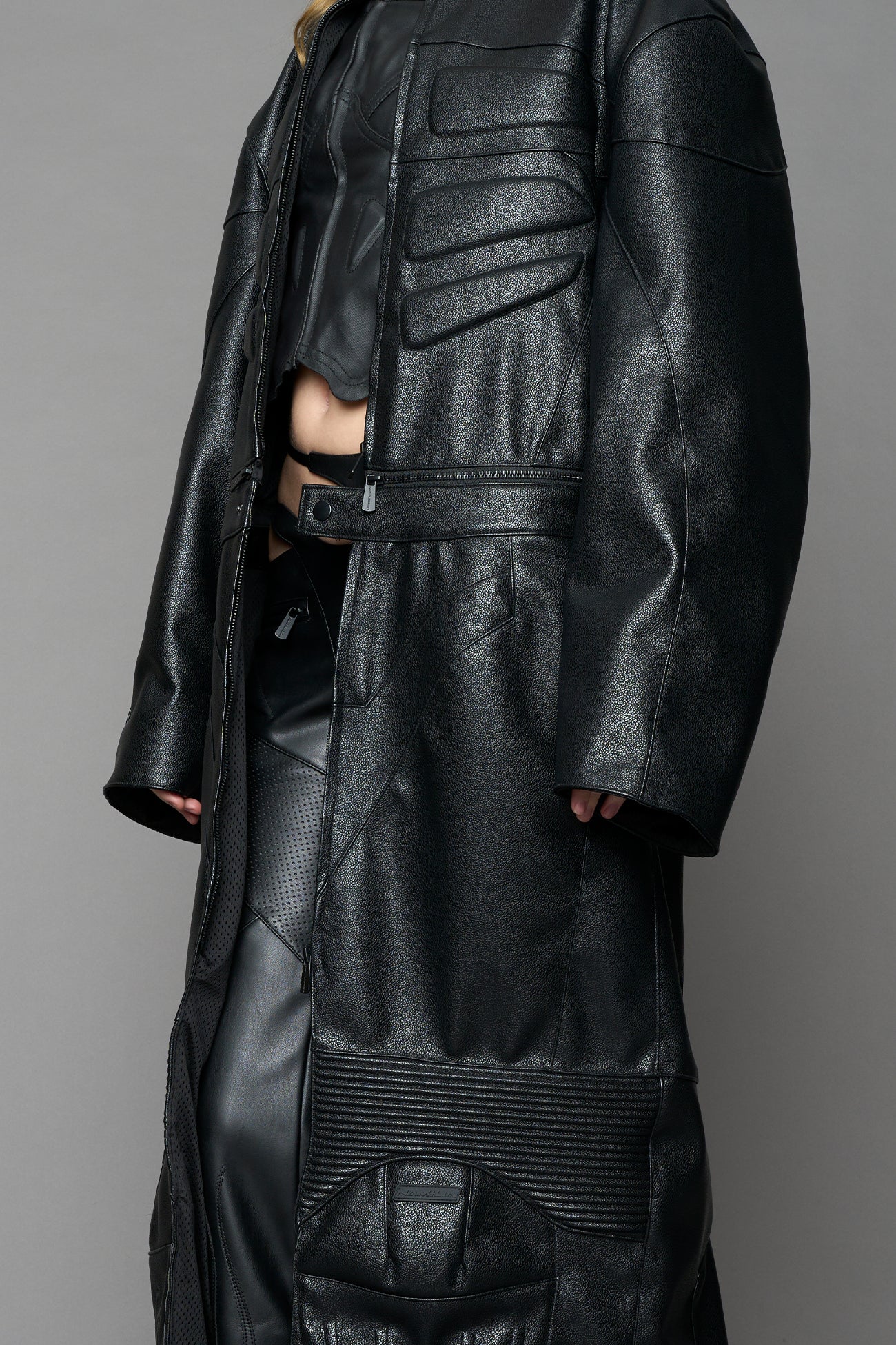 Detachable moto long coat in black vegan black leather inspired by a deconstructed full-body motocross racing suit, featuring padded panel details and a detachable zip design for two-way wear.