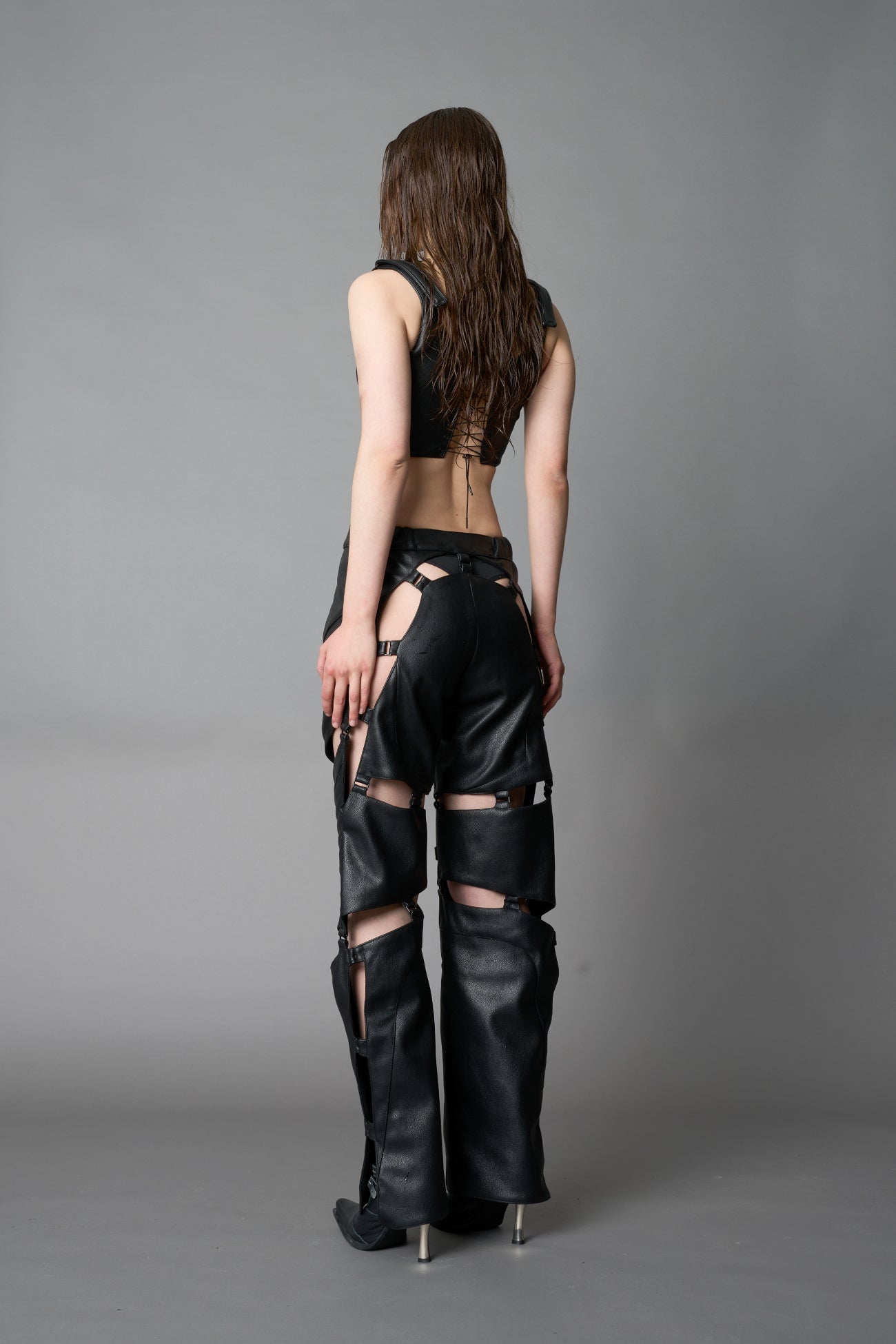female Model wearing black vegan leather motocross-inspired pants with thigh cut-outs, strap harness details, quilted knee padding, and full-length leg gaiters; styled with a structured black bandeau top against a neutral background.