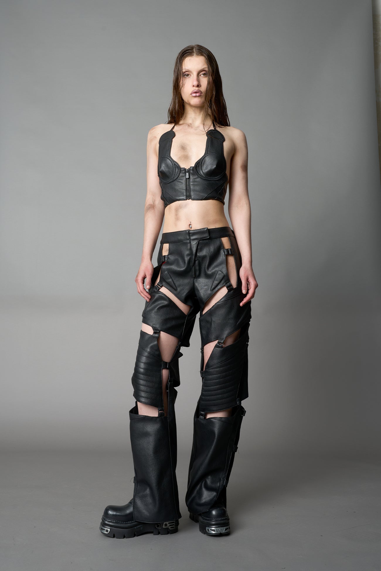 female Model wearing black vegan leather motocross-inspired pants with thigh cut-outs, strap harness details, quilted knee padding, and full-length leg gaiters; styled with a structured black bandeau top against a neutral background.