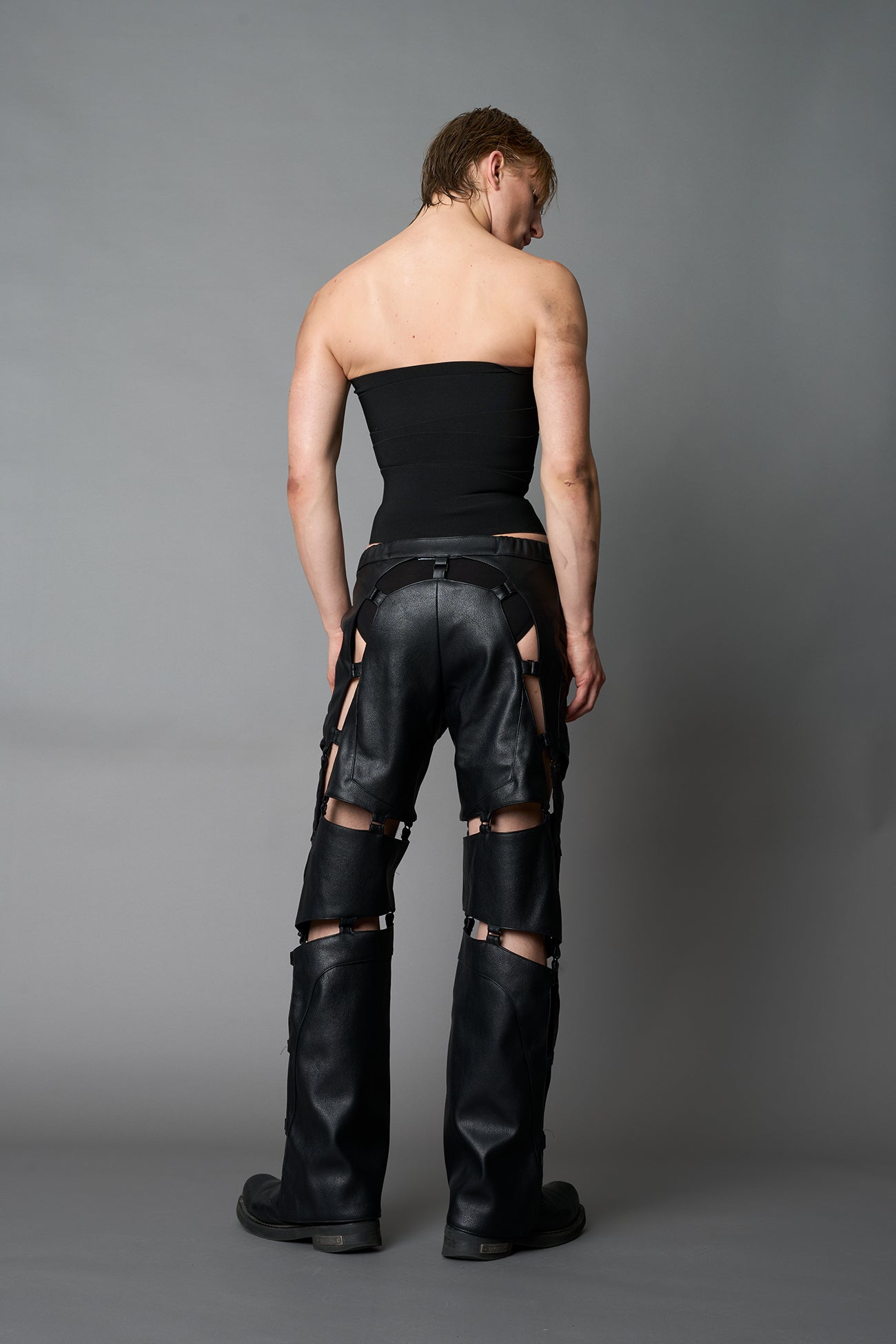 male Model wearing black vegan leather motocross-inspired pants with thigh cut-outs, strap harness details, quilted knee padding, and full-length leg gaiters; styled with a structured black bandeau top against a neutral background.