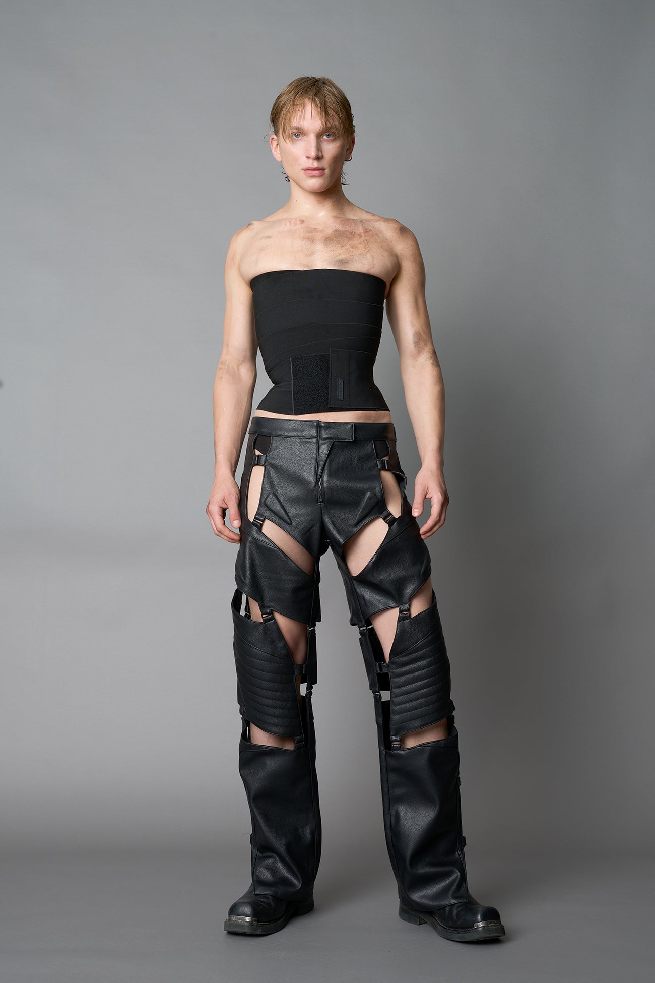 male Model wearing black vegan leather motocross-inspired pants with thigh cut-outs, strap harness details, quilted knee padding, and full-length leg gaiters; styled with a structured black bandeau top against a neutral background.