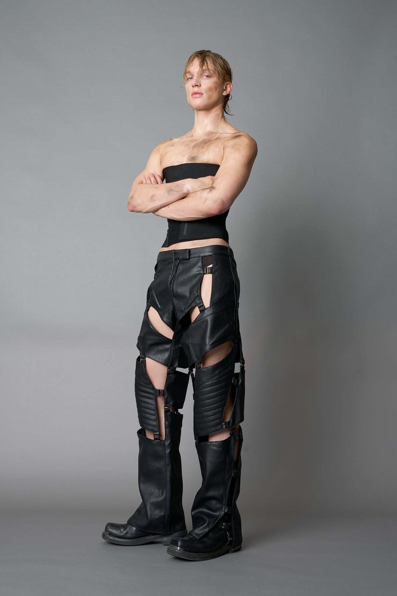 male Model wearing black vegan leather motocross-inspired pants with thigh cut-outs, strap harness details, quilted knee padding, and full-length leg gaiters; styled with a structured black bandeau top against a neutral background.