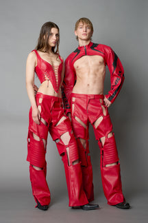 Model wearing red vegan leather motocross-inspired pants with thigh cut-outs, strap harness details, quilted knee padding, and full-length leg gaiters; styled with a structured black bandeau top against a neutral background.