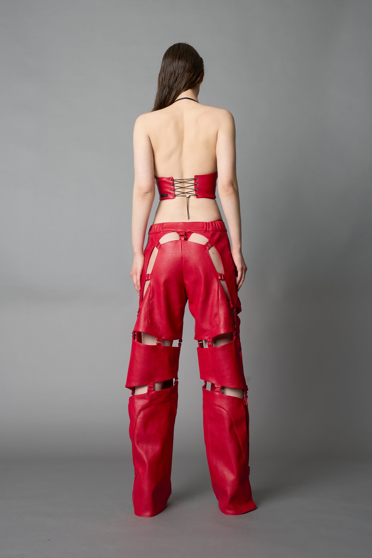Model wearing red vegan leather motocross-inspired pants with thigh cut-outs, strap harness details, quilted knee padding, and full-length leg gaiters; styled with a structured black bandeau top against a neutral background.