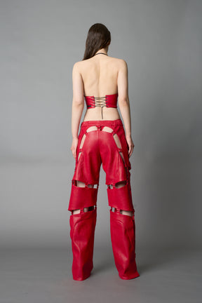 Model wearing red vegan leather motocross-inspired pants with thigh cut-outs, strap harness details, quilted knee padding, and full-length leg gaiters; styled with a structured black bandeau top against a neutral background.