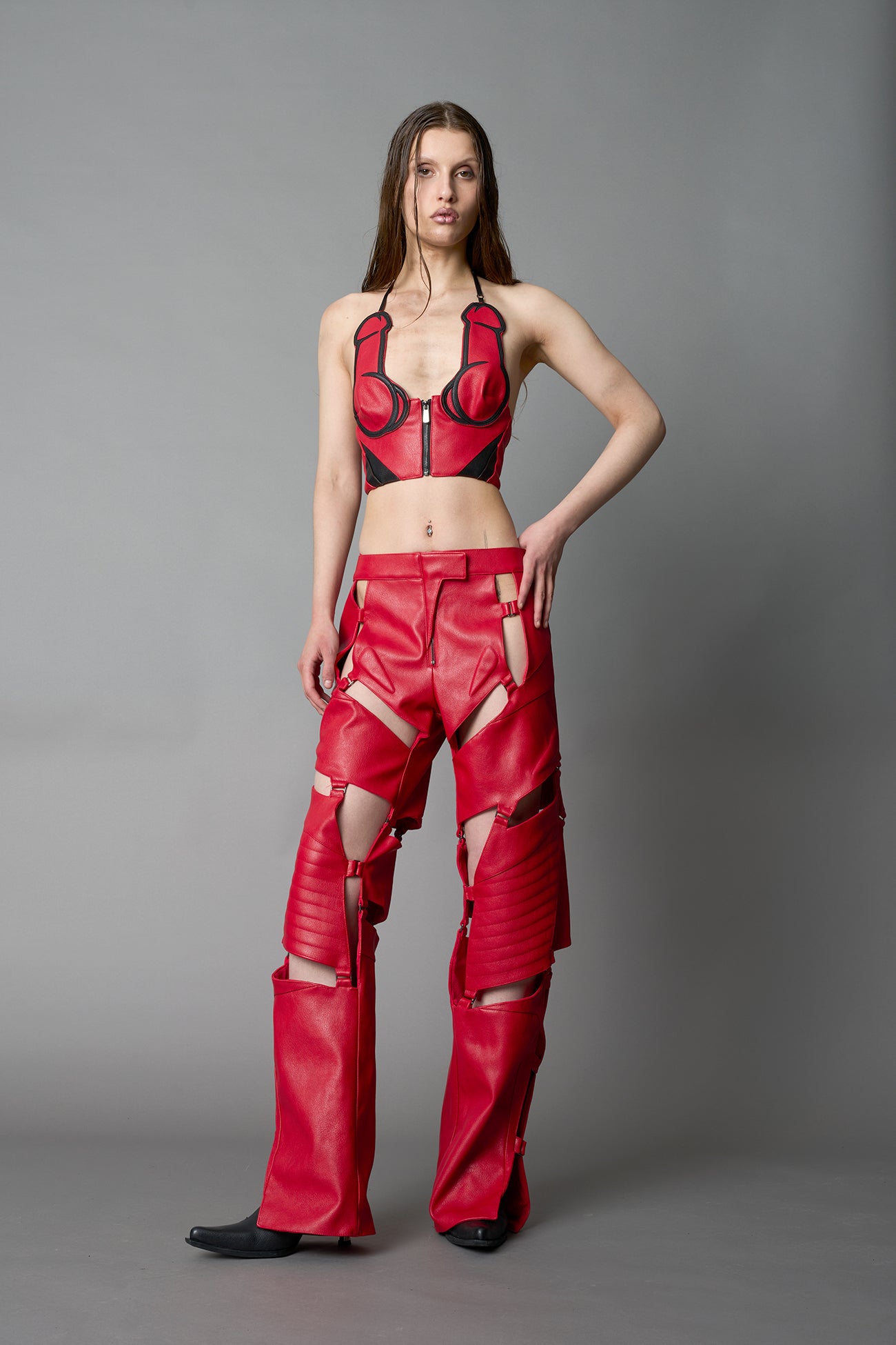 Model wearing red vegan leather motocross-inspired pants with thigh cut-outs, strap harness details, quilted knee padding, and full-length leg gaiters; styled with a structured black bandeau top against a neutral background.