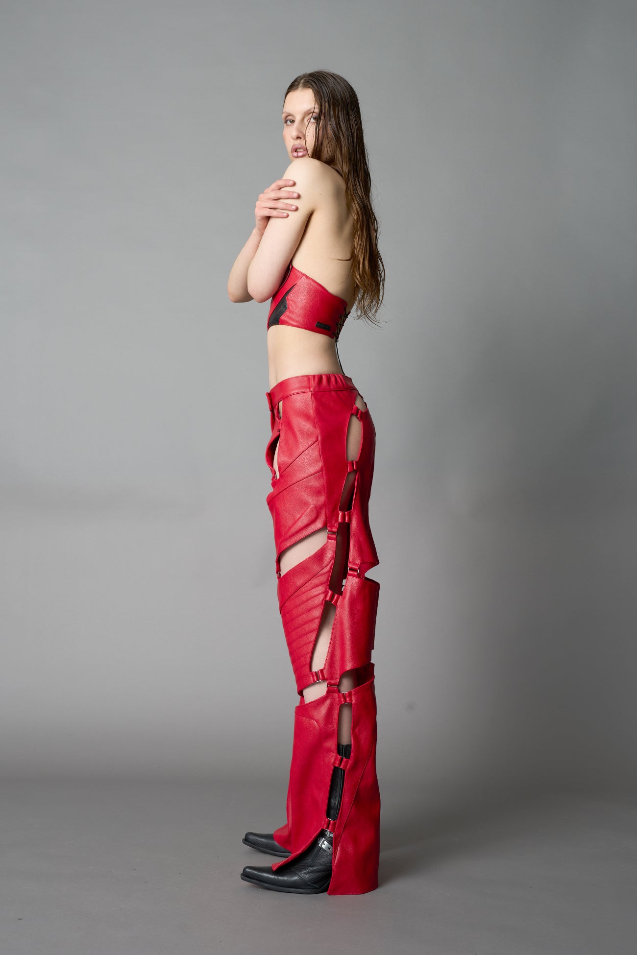 Model wearing red vegan leather motocross-inspired pants with thigh cut-outs, strap harness details, quilted knee padding, and full-length leg gaiters; styled with a structured black bandeau top against a neutral background.