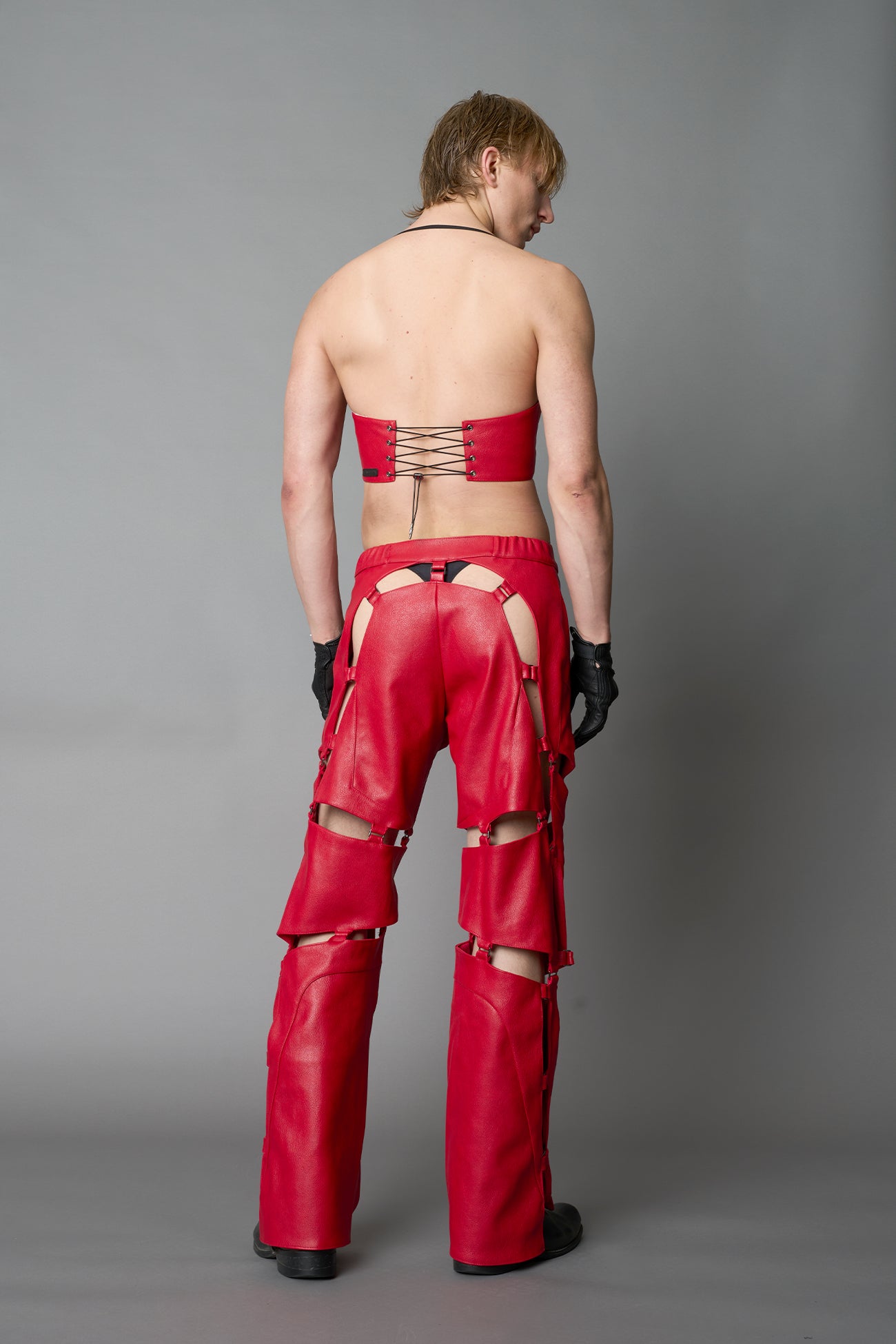 Model wearing red vegan leather motocross-inspired pants with thigh cut-outs, strap harness details, quilted knee padding, and full-length leg gaiters; styled with a structured black bandeau top against a neutral background.