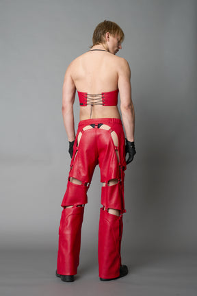 Model wearing red vegan leather motocross-inspired pants with thigh cut-outs, strap harness details, quilted knee padding, and full-length leg gaiters; styled with a structured black bandeau top against a neutral background.