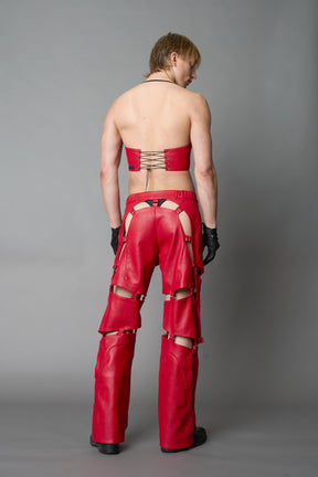 Model wearing red vegan leather motocross-inspired pants with thigh cut-outs, strap harness details, quilted knee padding, and full-length leg gaiters; styled with a structured black bandeau top against a neutral background.