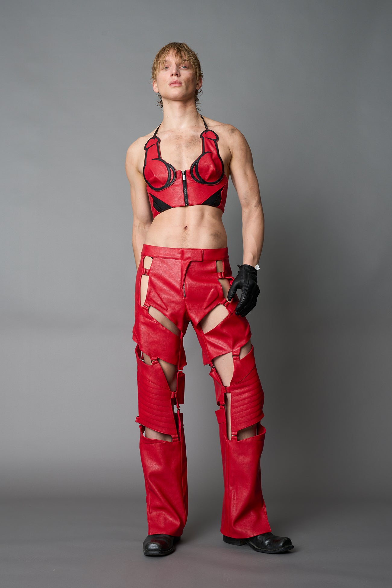 Model wearing red vegan leather motocross-inspired pants with thigh cut-outs, strap harness details, quilted knee padding, and full-length leg gaiters; styled with a structured black bandeau top against a neutral background.