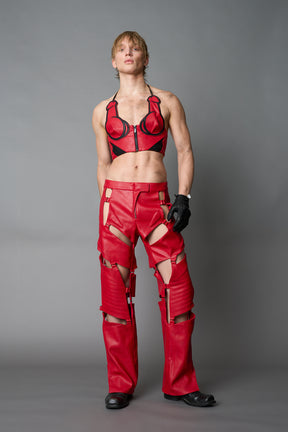 Model wearing red vegan leather motocross-inspired pants with thigh cut-outs, strap harness details, quilted knee padding, and full-length leg gaiters; styled with a structured black bandeau top against a neutral background.