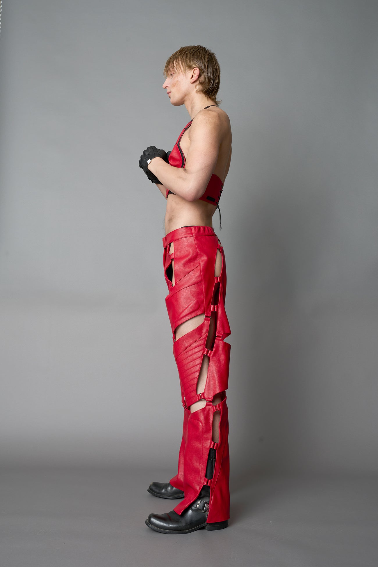Model wearing red vegan leather motocross-inspired pants with thigh cut-outs, strap harness details, quilted knee padding, and full-length leg gaiters; styled with a structured black bandeau top against a neutral background.
