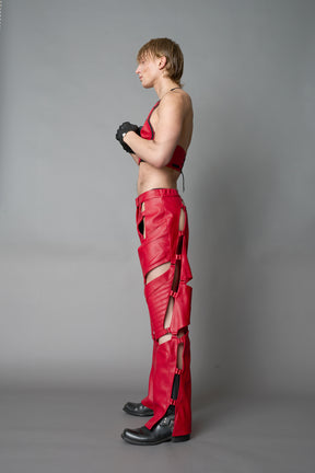 Model wearing red vegan leather motocross-inspired pants with thigh cut-outs, strap harness details, quilted knee padding, and full-length leg gaiters; styled with a structured black bandeau top against a neutral background.