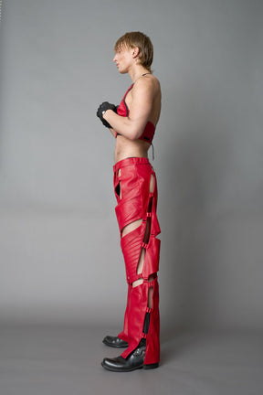 Model wearing red vegan leather motocross-inspired pants with thigh cut-outs, strap harness details, quilted knee padding, and full-length leg gaiters; styled with a structured black bandeau top against a neutral background.