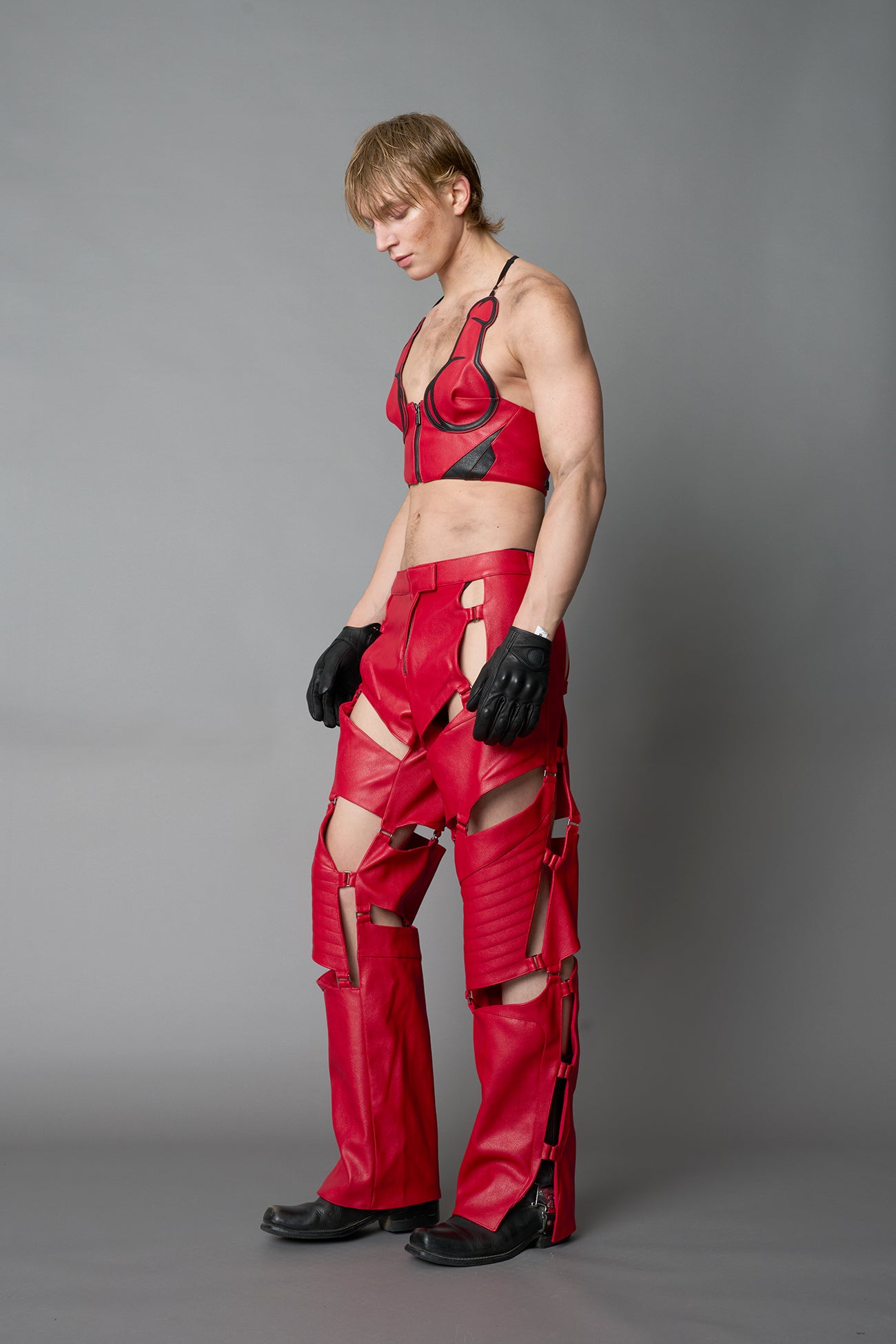 Model wearing red vegan leather motocross-inspired pants with thigh cut-outs, strap harness details, quilted knee padding, and full-length leg gaiters; styled with a structured black bandeau top against a neutral background.