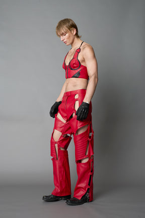 Model wearing red vegan leather motocross-inspired pants with thigh cut-outs, strap harness details, quilted knee padding, and full-length leg gaiters; styled with a structured black bandeau top against a neutral background.