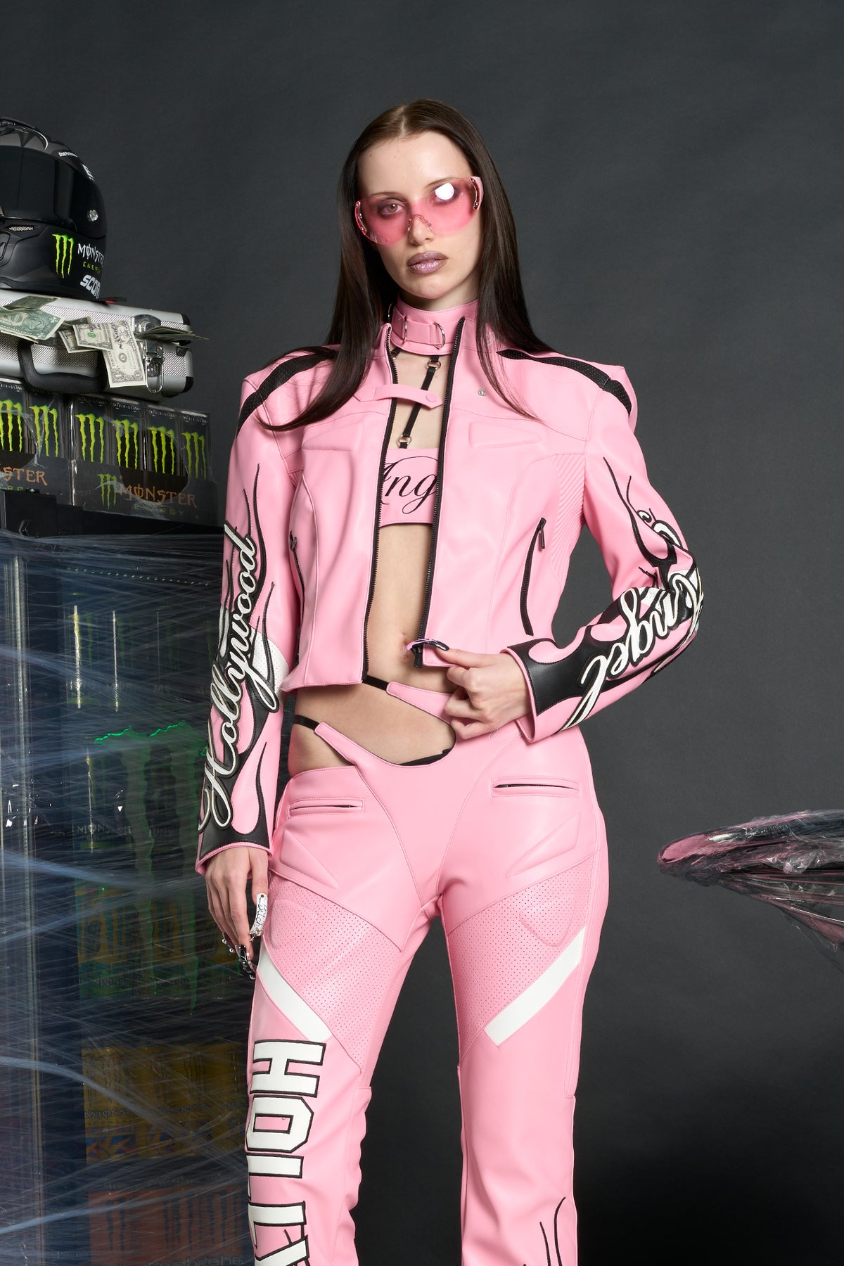 Model wears a pink motocross-style jacket made from vegan PU leather with flame topstitching, padded perforated paneling, topstiched sleeve slogan, ‘Angel’ embroidery on collar, and branded zippered front and pockets.