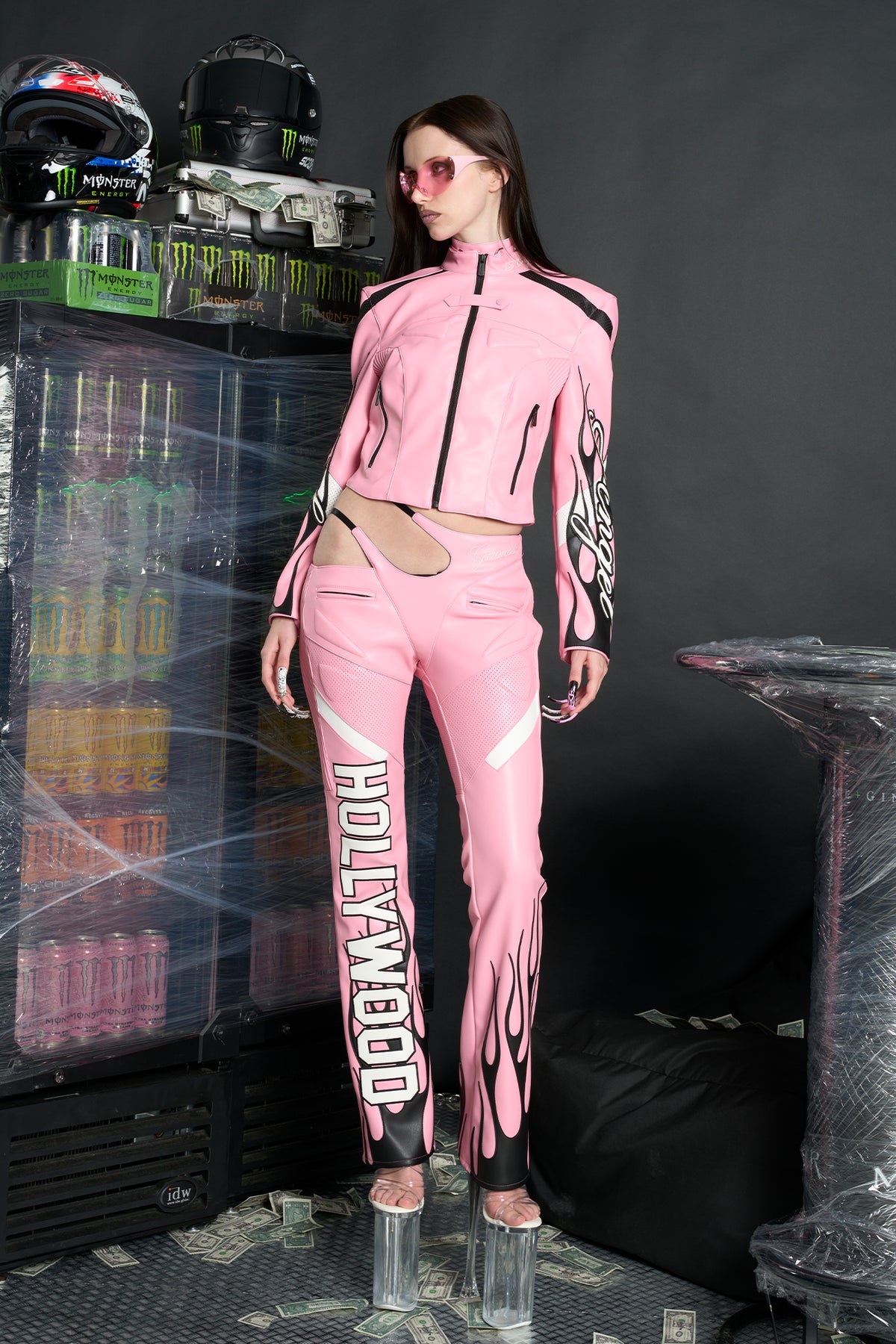Model wears a pink motocross-style jacket made from vegan PU leather with flame topstitching, padded perforated paneling, topstiched sleeve slogan, ‘Angel’ embroidery on collar, and branded zippered front and pockets.