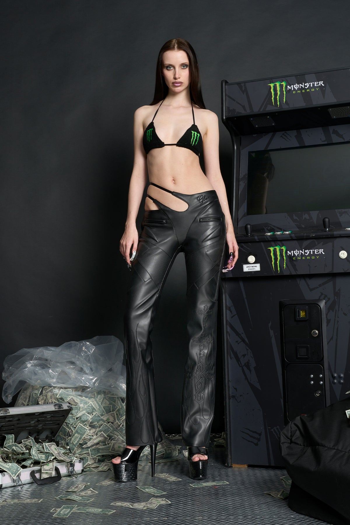 Model wears black vegan leather flare trousers with motocross paneling, padded details, flame and cross stitching, slogan embroidery, and an asymmetric waistband with adjustable elastic thong straps.