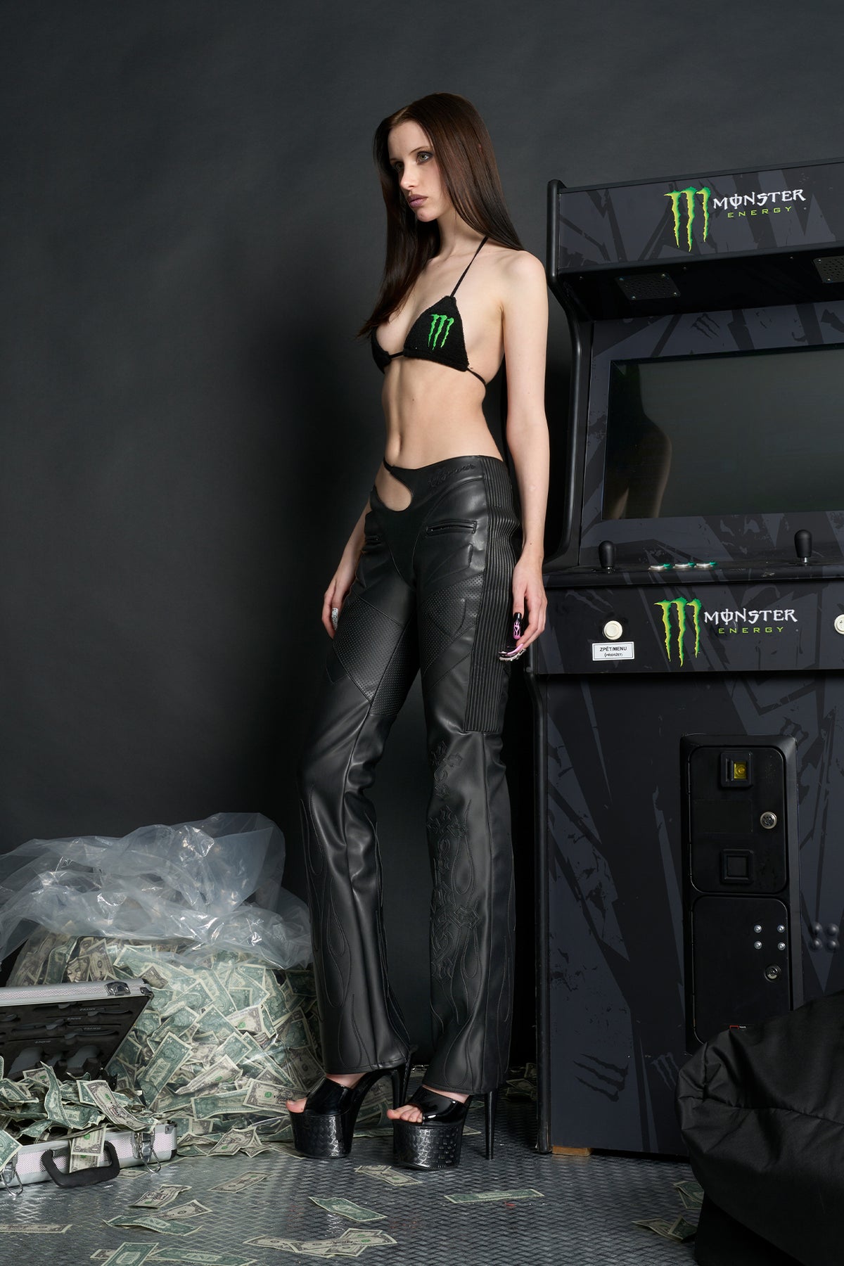 Model wears black vegan leather flare trousers with motocross paneling, padded details, flame and cross stitching, slogan embroidery, and an asymmetric waistband with adjustable elastic thong straps.