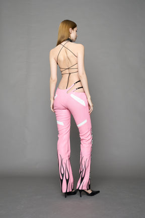 Model wears pink vegan leather flare trousers with motocross paneling, padded details, flame and cross stitching, slogan embroidery, and an asymmetric waistband with adjustable elastic thong straps.