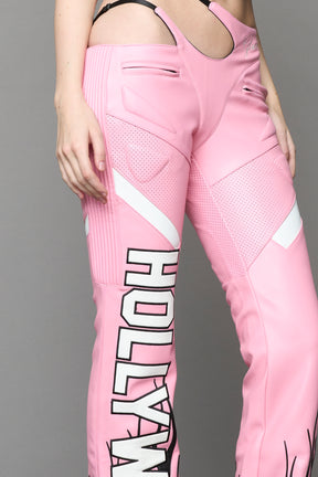 Model wears pink vegan leather flare trousers with motocross paneling, padded details, flame and cross stitching, slogan embroidery, and an asymmetric waistband with adjustable elastic thong straps.