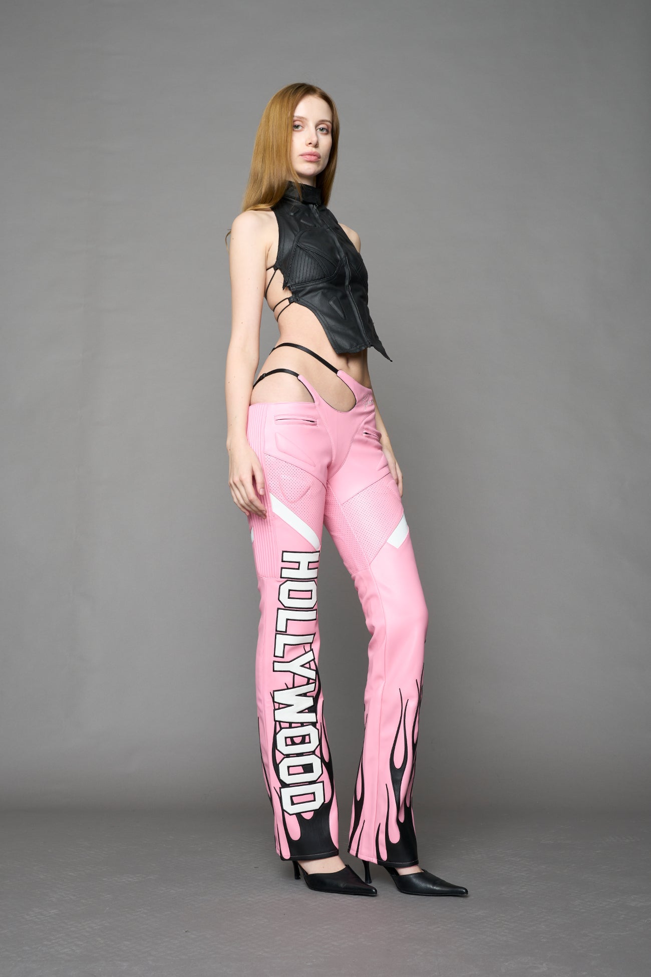 Model wears pink vegan leather flare trousers with motocross paneling, padded details, flame and cross stitching, slogan embroidery, and an asymmetric waistband with adjustable elastic thong straps.