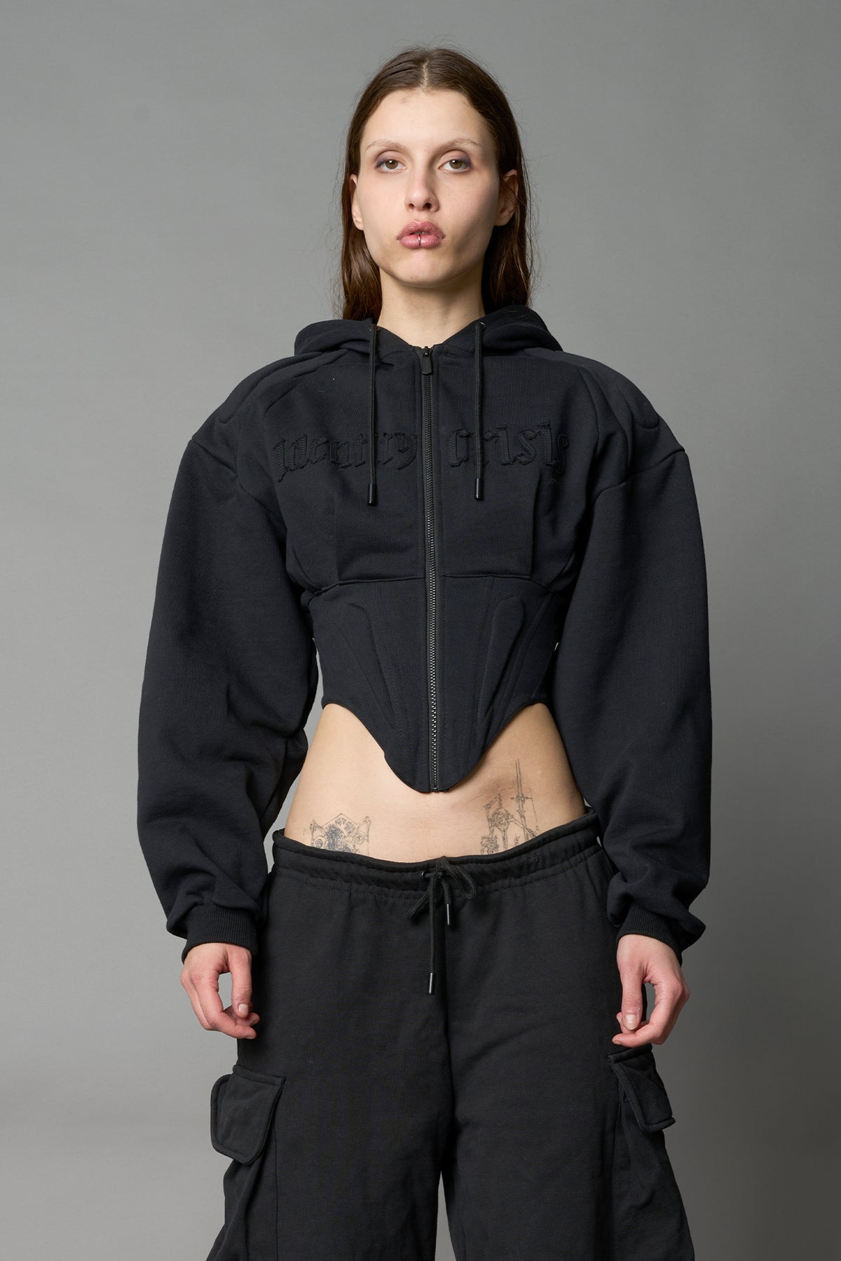 Front view of model in a black cropped hoodie featuring structured corset-style paneling  embroidered across the chest in tonal black