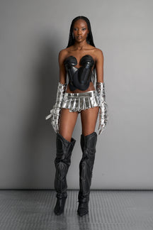 model wears the silver vegan PU leather Namilia Pleated Mini Skirt with ring piercing details on the hemline and velcro fold-over waistband closure.