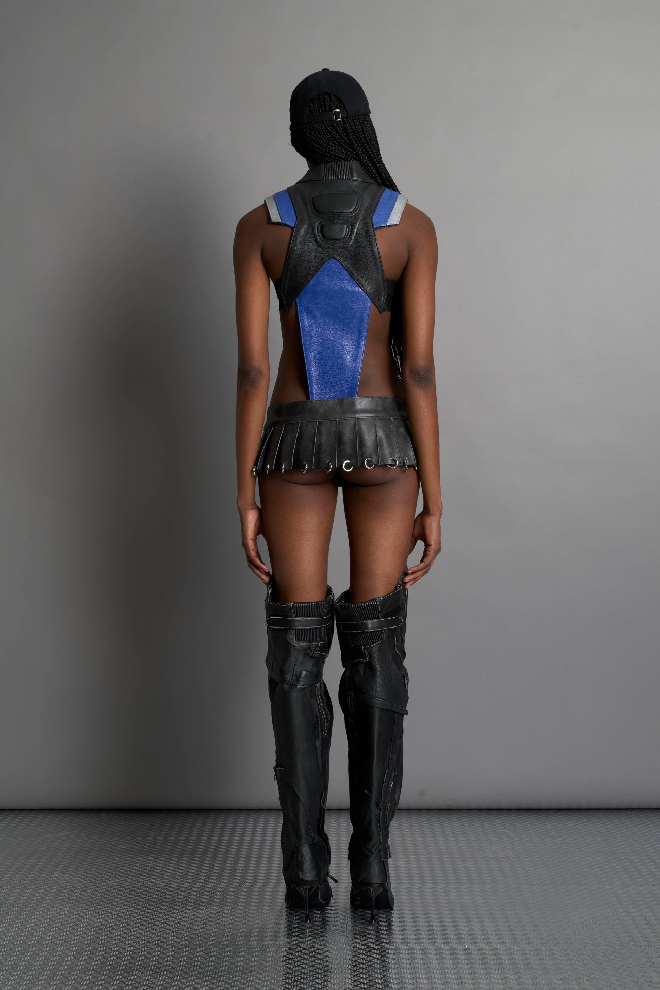 model wears the washed black vegan PU leather Namilia Pleated Mini Skirt with ring piercing details on the hemline and velcro fold-over waistband closure.