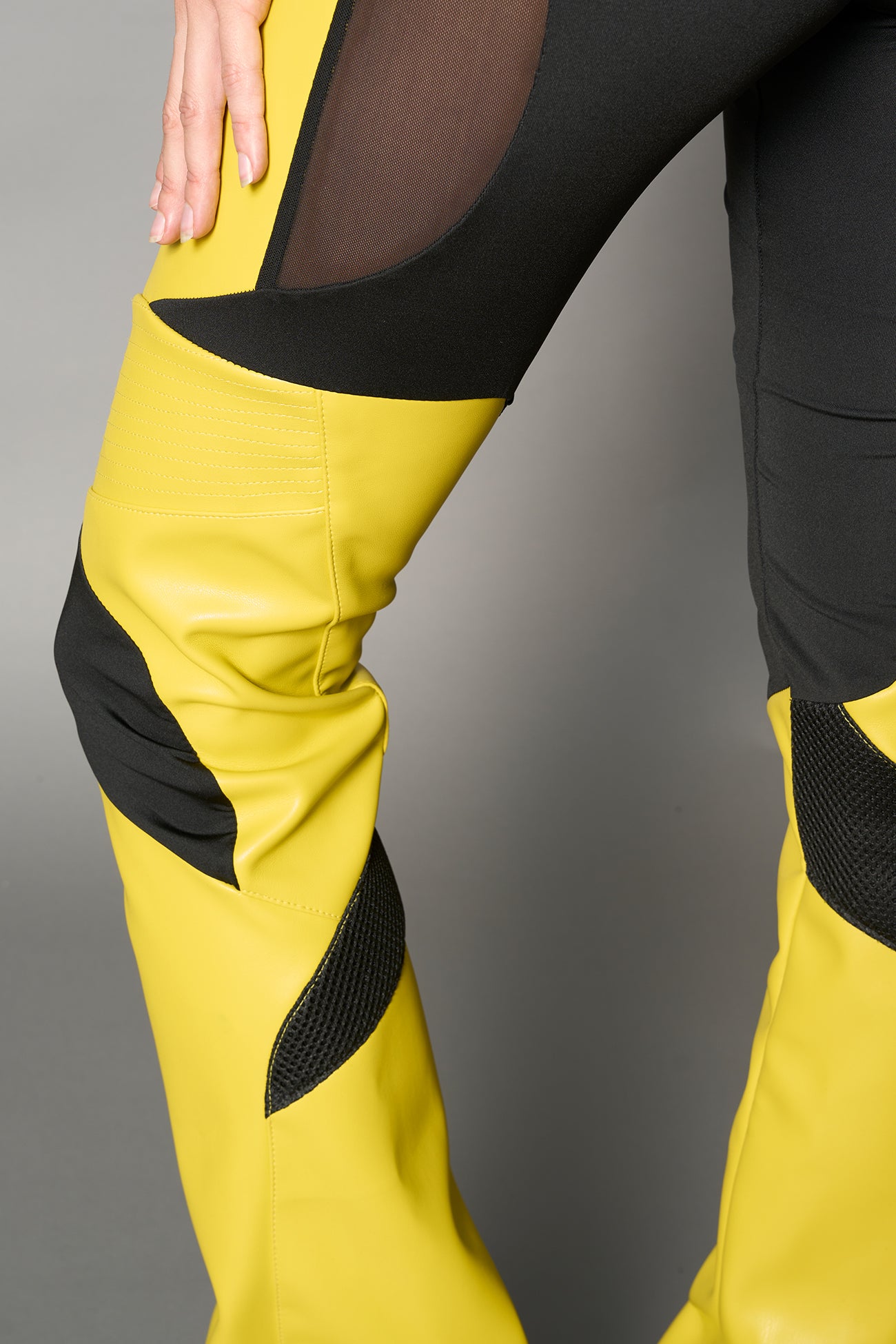 Model wearing high-waisted yellow vegan leather pants with mesh cutouts, moto-inspired paneling, and flared stacked legs – edgy streetwear trousers styled with chunky platform boots