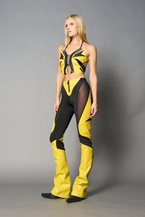 Model wearing high-waisted yellow vegan leather pants with mesh cutouts, moto-inspired paneling, and flared stacked legs – edgy streetwear trousers styled with chunky platform boots