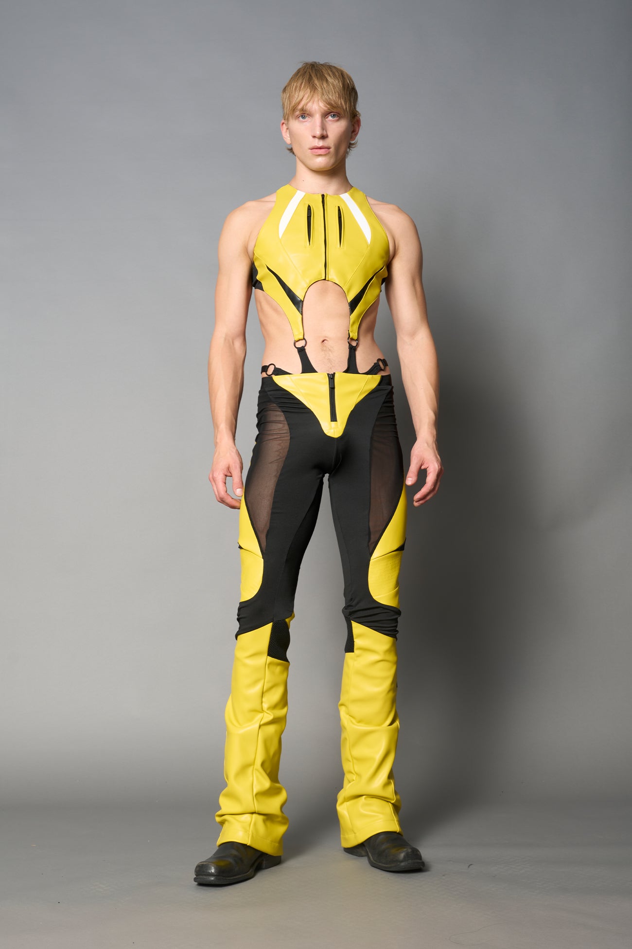 Model wearing high-waisted yellow vegan leather pants with mesh cutouts, moto-inspired paneling, and flared stacked legs – edgy streetwear trousers styled with chunky platform boots