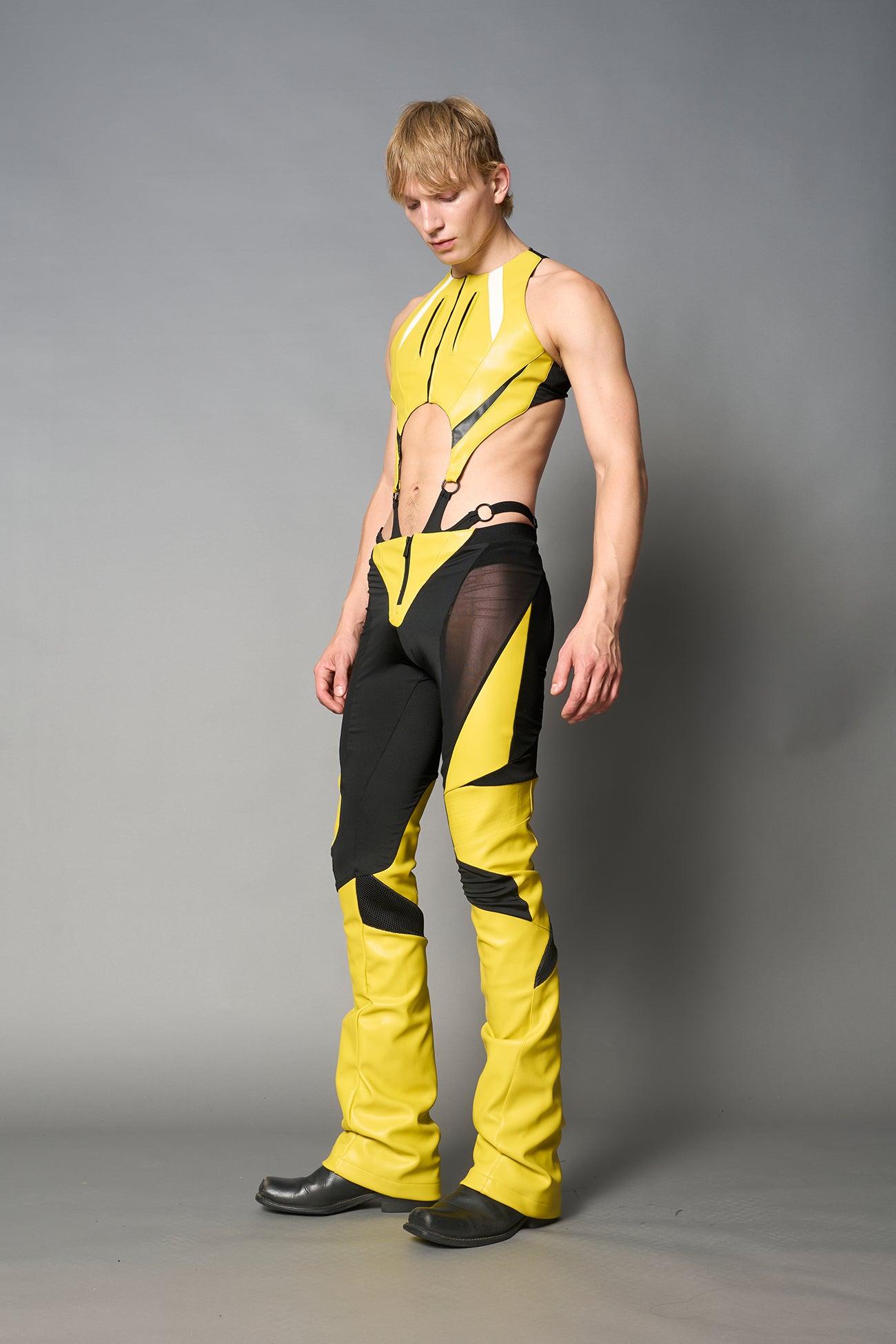 Model wearing high-waisted yellow vegan leather pants with mesh cutouts, moto-inspired paneling, and flared stacked legs – edgy streetwear trousers styled with chunky platform boots